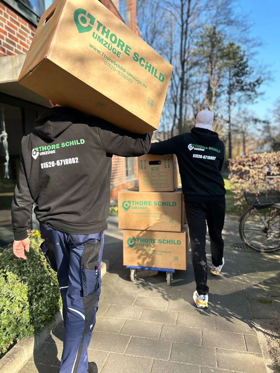 Zwei MItarbeiter von Thore Schild Umzüge transportieren mehrere Umzugskartons zu einem Haus.